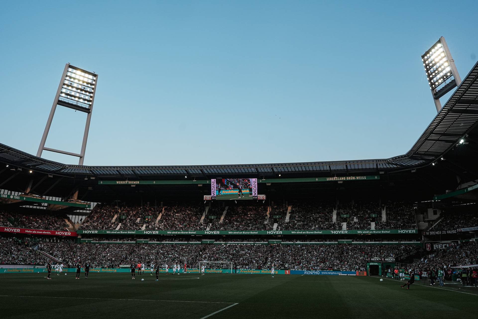 Ein großes, beleuchtetes Fußballstadion voller Zuschauer unter strahlend blauem Himmel während eines Spiels.