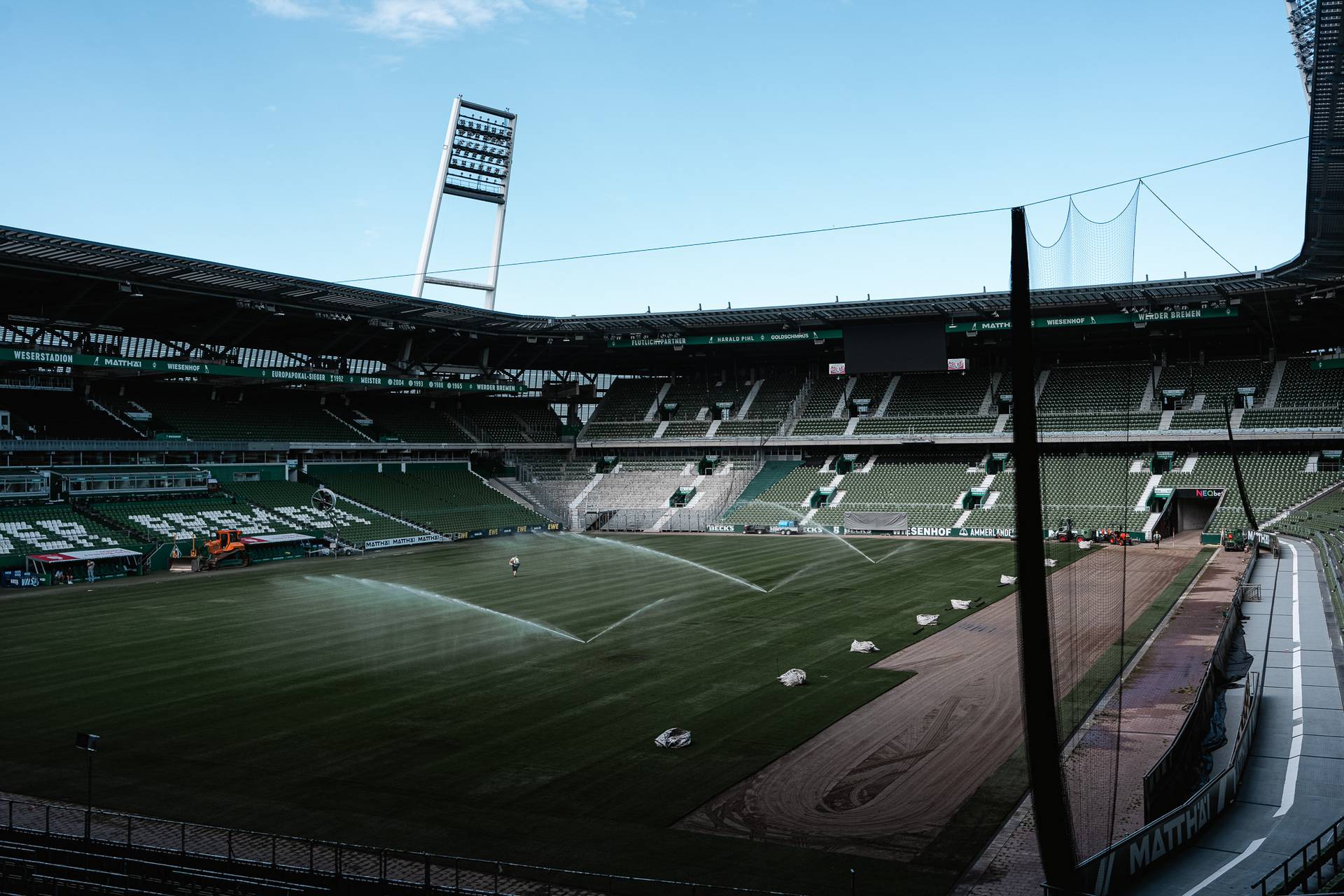 Ein leeres Fußballstadion mit grünen Sitzen und einem gepflegten Rasen, der gerade bewässert wird. Große Flutlichter und ein blauer Himmel sind zu sehen.