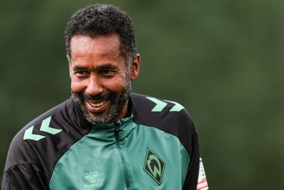 Ein lächelnder Mann mit lockigem Haar, der eine grün-schwarze Sportjacke mit Logo trägt und im Freien vor einem unscharfen grünen Hintergrund steht.
