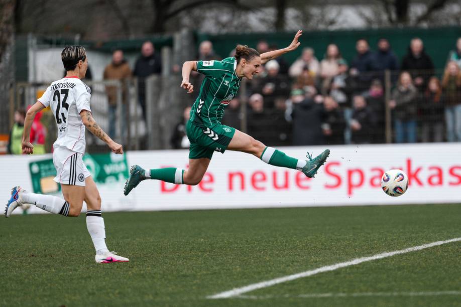 Zwei Fußballspieler auf dem Spielfeld; einer in Grün, der gerade in der Luft einen Schuss abgibt, und ein anderer in Weiß, der hinter ihm herläuft. Im Hintergrund sind Zuschauer und eine digitale Anzeigetafel zu sehen.