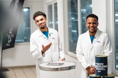 Justin Njinmah und Felix Agu lachen nebeneinander. Sie tragen lange weiße Arbeitskittel und stehen in einer Fabrik vor einem Pult.