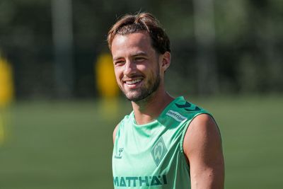 Ein lächelnder Mann in einem grünen Sporttrikot steht an einem sonnigen Tag im Freien, vor einem verschwommenen Hintergrund aus Grün und gelben Formen.