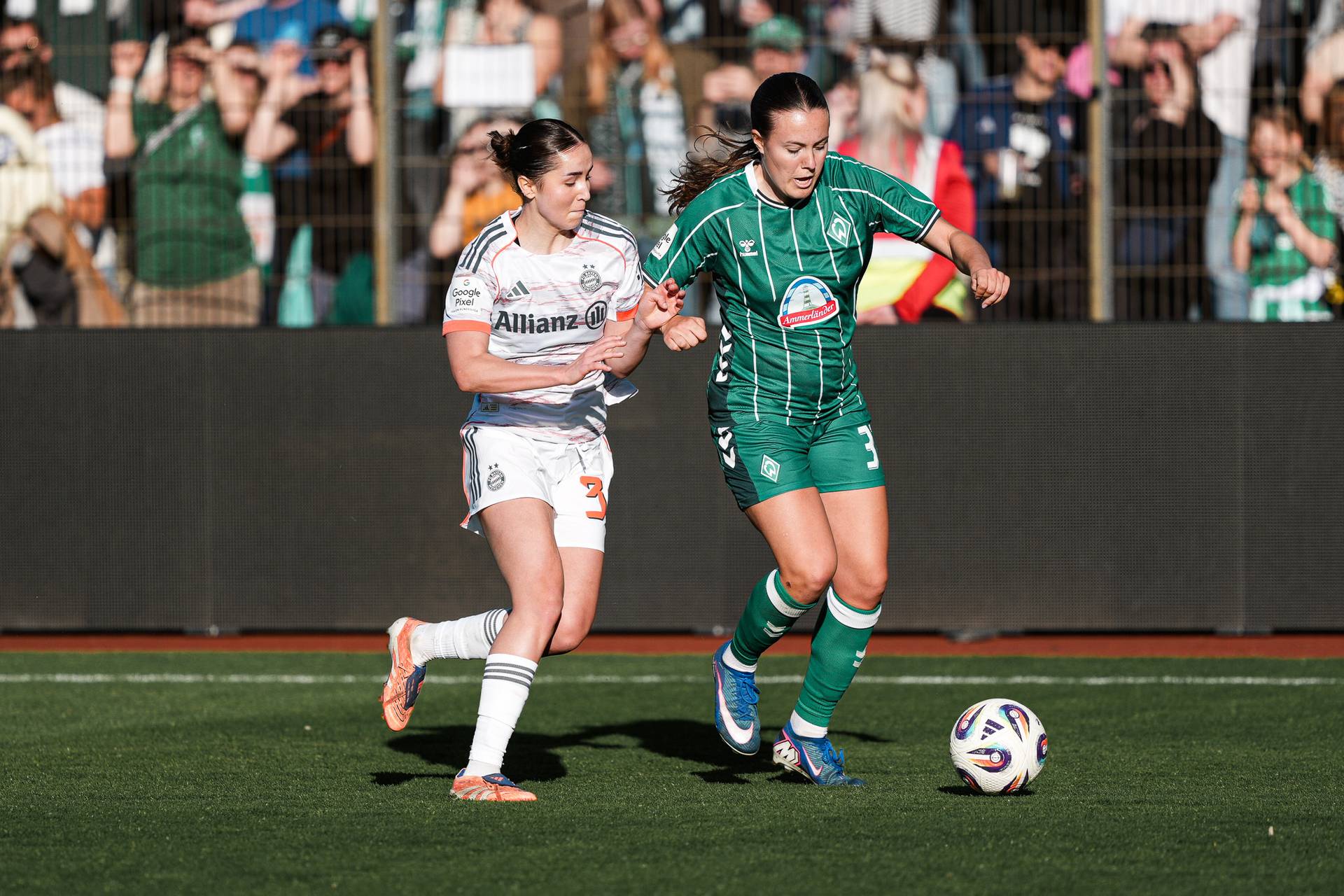 Zwei Fußballspielerinnen kämpfen auf einem sonnigen Spielfeld um den Ball, im Hintergrund sind Zuschauer zu sehen.