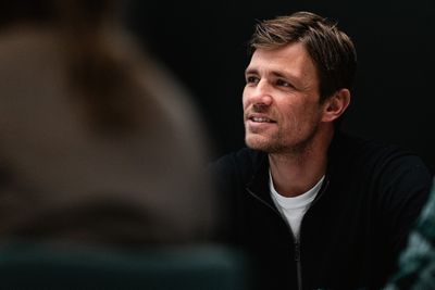 Ein Mann mit kurzen Haaren und Bart, der eine schwarze Jacke und ein weißes Hemd trägt, sitzt in einer schwach beleuchteten Umgebung.
