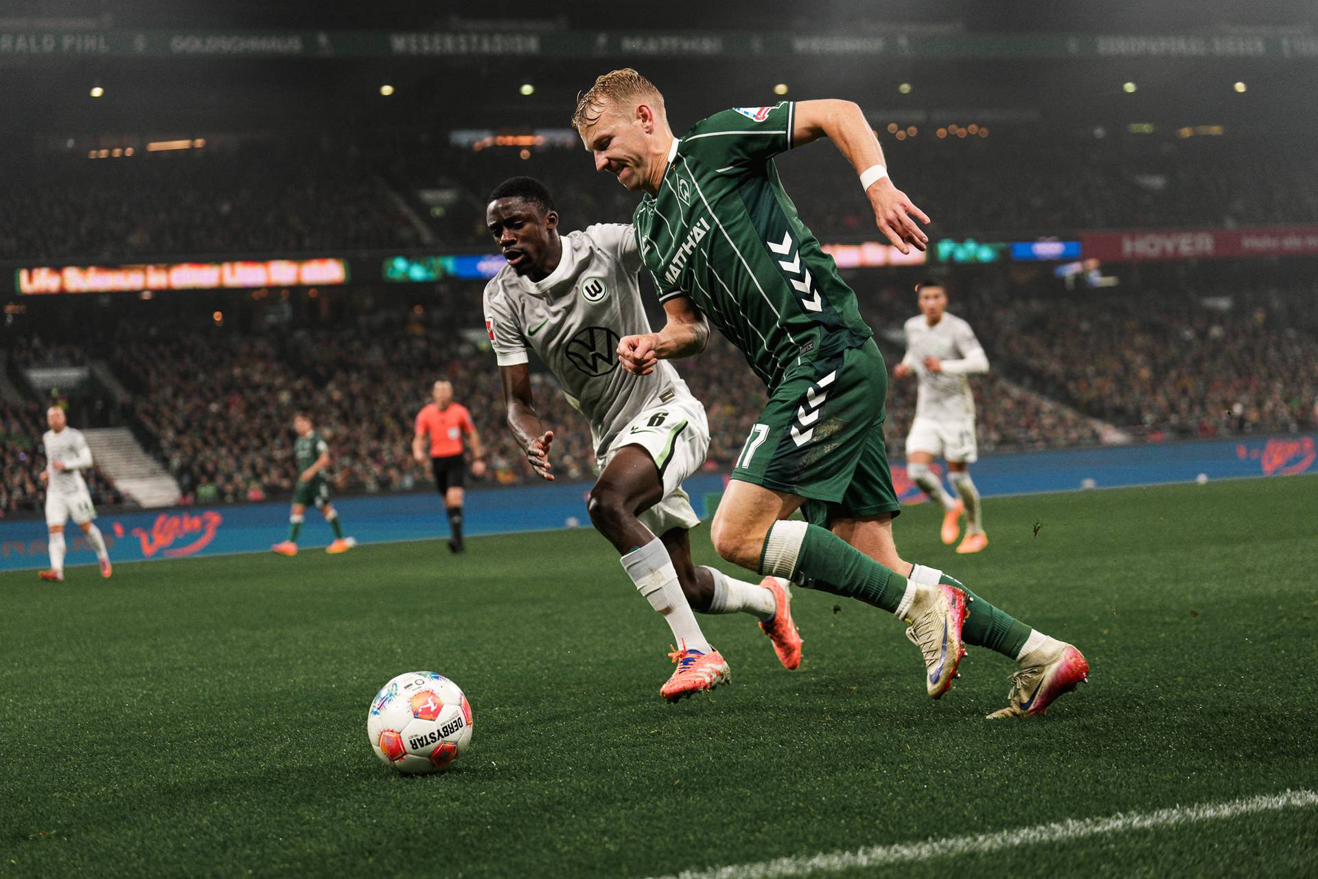 Zwei Fußballspieler, einer in Weiß und einer in Grün, kämpfen während eines Spiels auf dem Stadionrasen um den Ball.