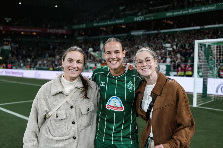 Michaela Brandenburg, Saskia Matheis und Hanna Nemeth lächeln auf einem Fußballplatz, eine von ihnen trägt ein grünes Trikot von Werder Bremen, im Hintergrund ist eine Menschenmenge zu sehen.