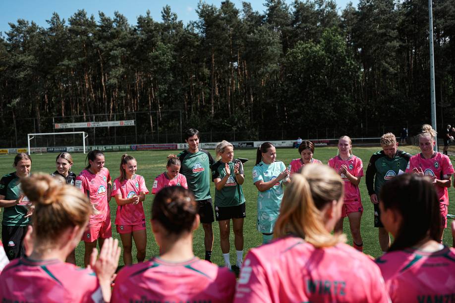 Eine Gruppe von Fußballspielern in korallfarbenen und grünen Trikots steht im Halbkreis auf einem Rasenfeld, klatscht und lächelt, im Hintergrund ist eine Baumreihe zu sehen.