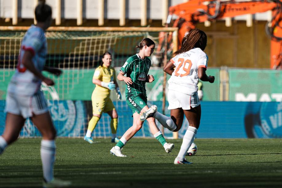 Fußballspielerin in Aktion auf dem Spielfeld: Einer in Grün dribbelt den Ball an einem Spieler in Weiß vorbei, während ein Torwart in Gelb bereit steht.