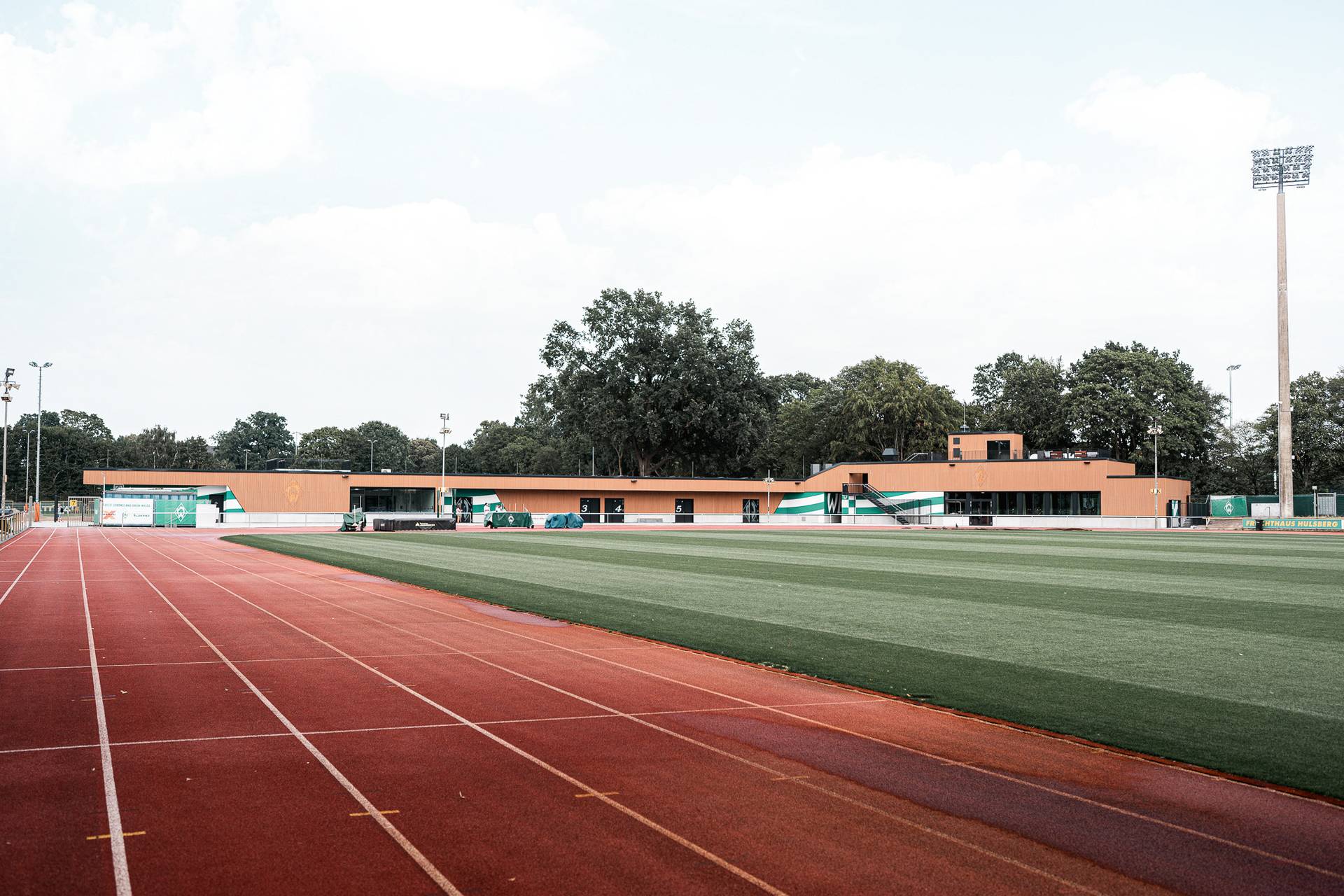 Eine Laufbahn führt zu einer Sportanlage mit einem Holzgebäude, einer grünen Wiese, Bäumen und Flutlicht unter einem bewölkten Himmel.