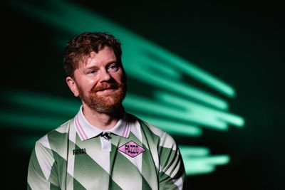 Ein lächelnder Mann mit Bart, der ein grün-weiß gemustertes Hemd trägt, vor einem dunklen Hintergrund mit grünen Lichtstreifen.
