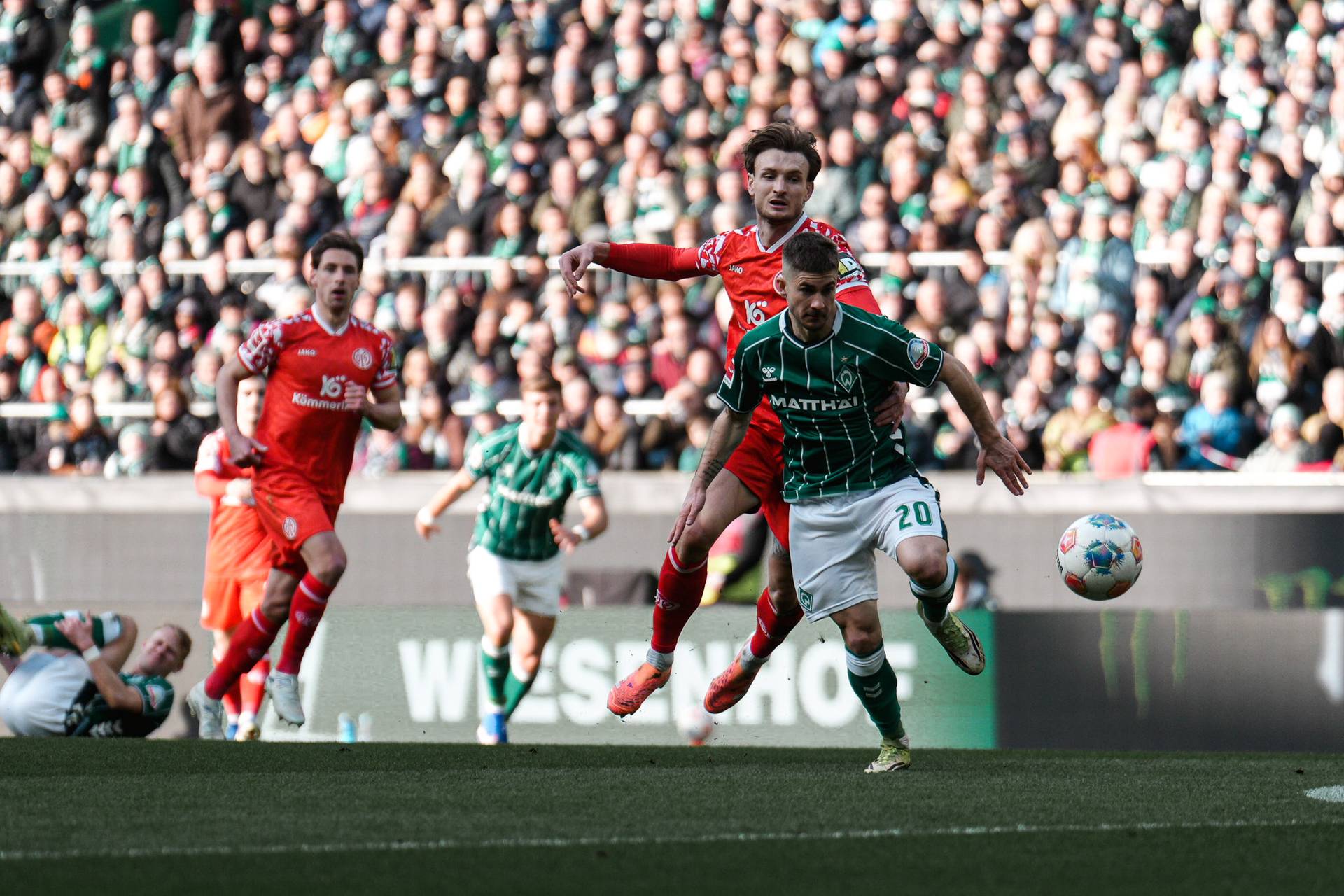 Fußballspieler in Aktion während eines Spiels; einer in Grün läuft mit dem Ball vorwärts, umgeben von gegnerischen Spielern in Rot, im Hintergrund ein vollbesetztes Stadion.
