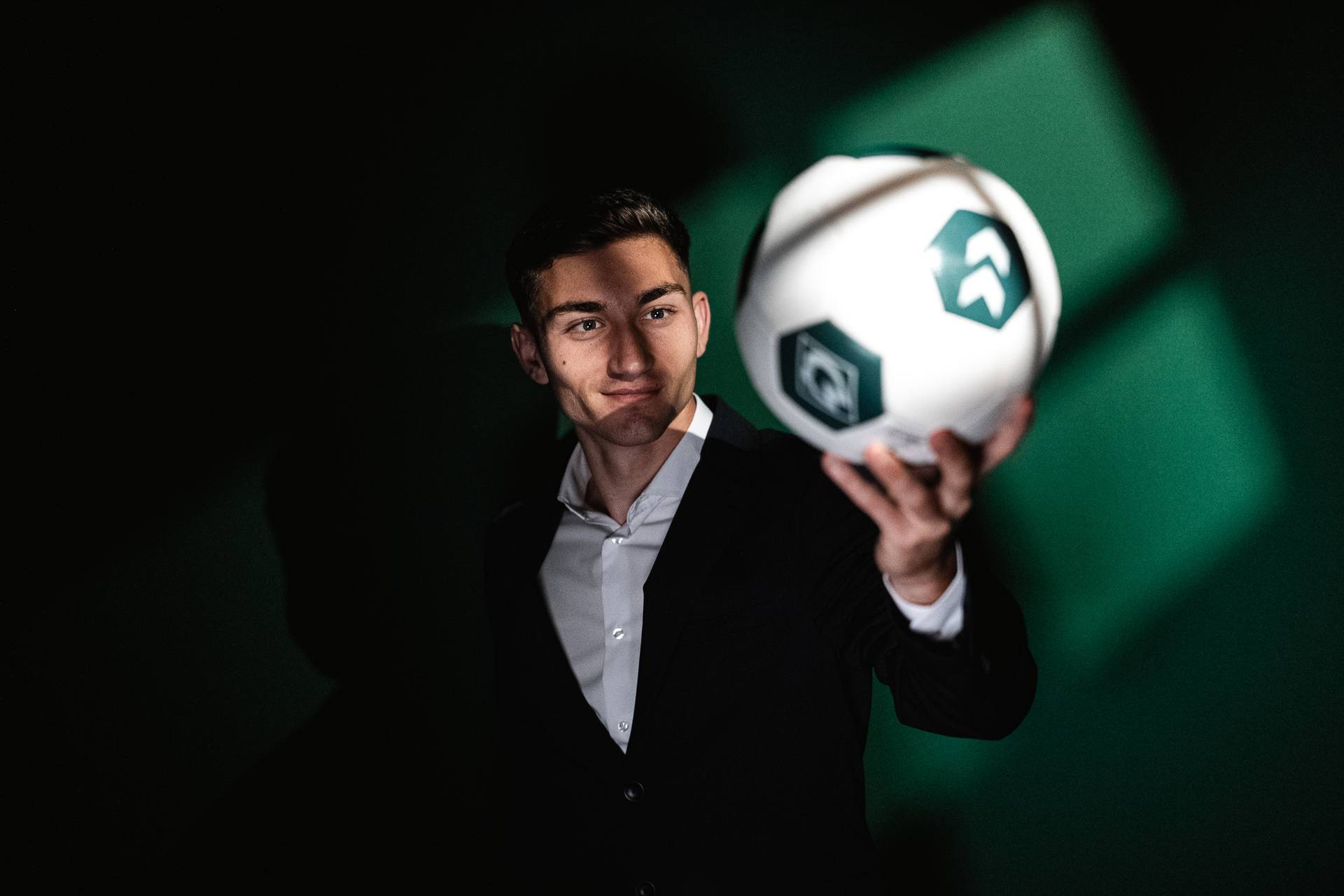 Eine Person im Anzug hält selbstbewusst einen Fußball mit grün-weißen Feldern in der Hand, vor einem dunklen Hintergrund mit Licht.