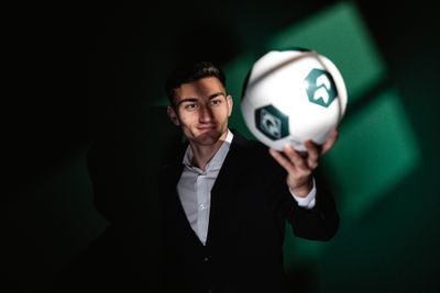 Eine Person im Anzug hält selbstbewusst einen Fußball mit grün-weißen Feldern in der Hand, vor einem dunklen Hintergrund mit Licht.