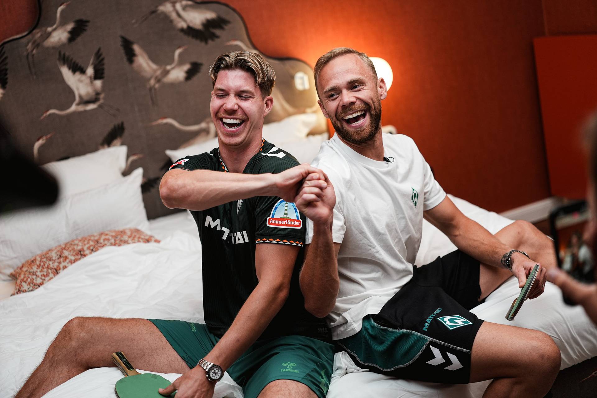 Zwei Männer sitzen auf einem Bett, lachen und stoßen mit den Fäusten aneinander, wobei jeder einen Tischtennisschläger in der Hand hält.