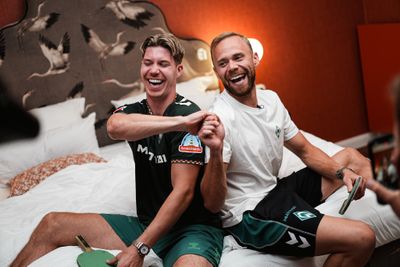 Zwei Männer sitzen auf einem Bett, lachen und stoßen mit den Fäusten aneinander, wobei jeder einen Tischtennisschläger in der Hand hält.