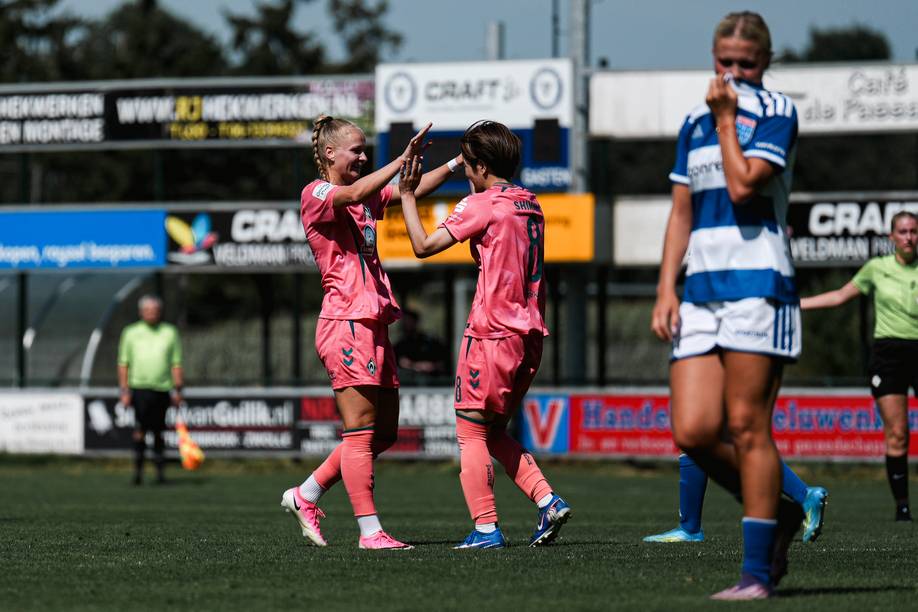 Zwei Fußballspielerinnen in korallroten Trikots feiern auf dem Spielfeld mit High-Fives, während eine Spielerin in einem blau-weißen Trikot vorbeigeht.