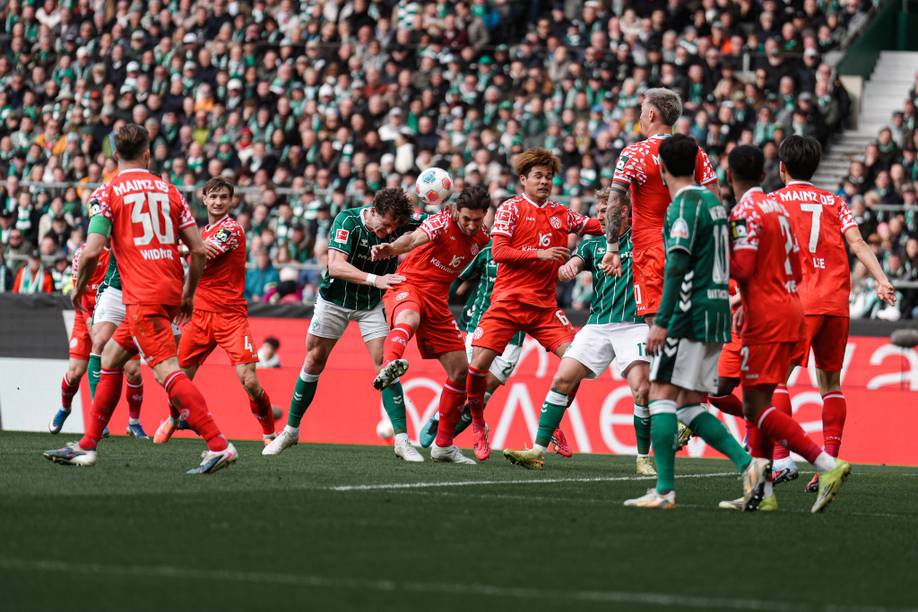 Fußballspieler in roten und grünen Trikots kämpfen in einem spannenden Spiel um den Ball, umgeben von einem vollbesetzten Stadion.