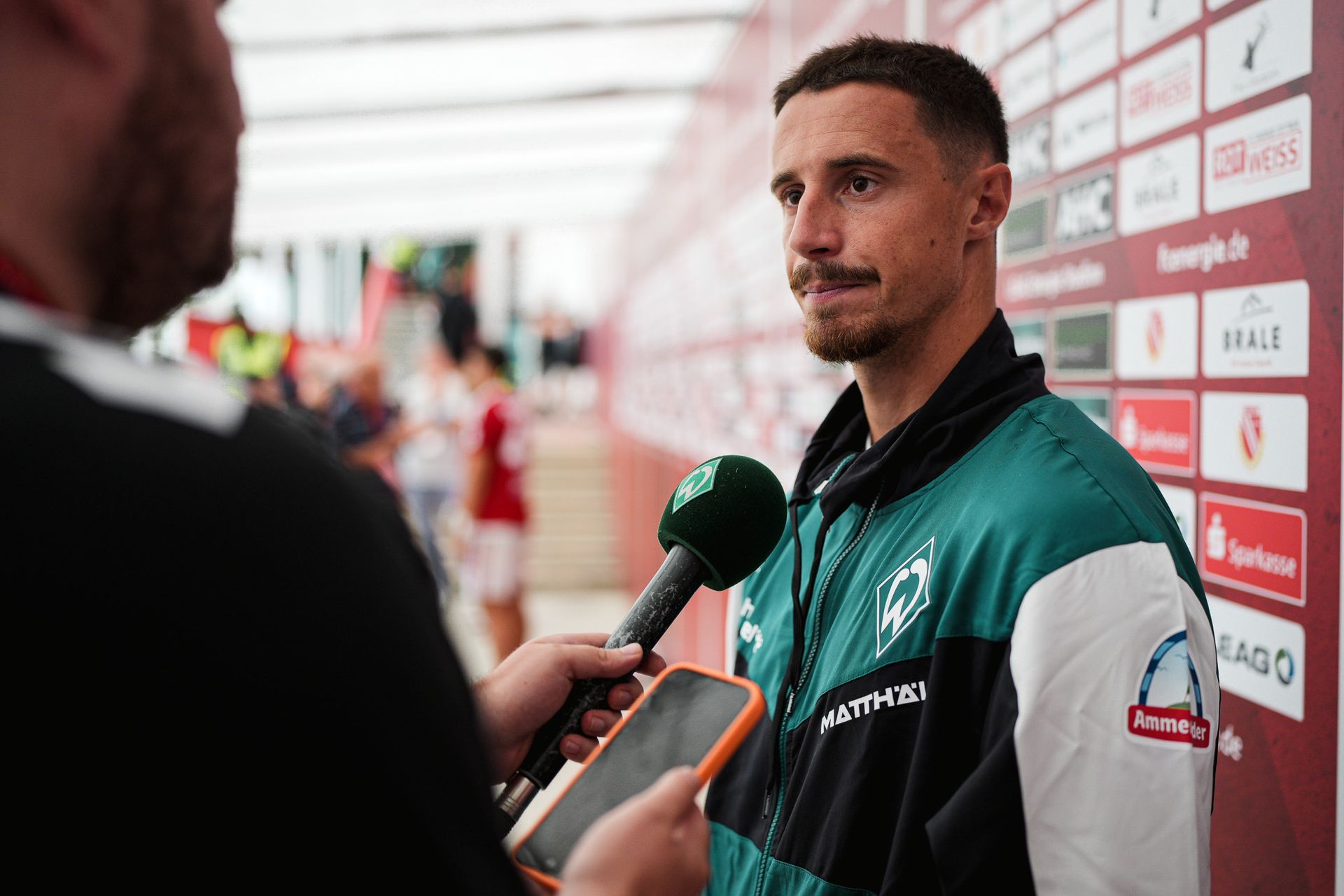 Ein Mann in einer grünen Sportjacke, der mit einem Mikrofon interviewt wird und vor einem rot-weißen Sponsorenhintergrund steht.