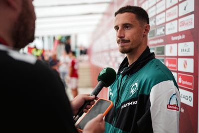 Ein Mann in einer grünen Sportjacke, der mit einem Mikrofon interviewt wird und vor einem rot-weißen Sponsorenhintergrund steht.