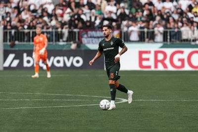 Ein Fußballspieler in einem schwarzen Trikot dribbelt den Ball auf einem Spielfeld, im Hintergrund sind ein Torwart in einem orangefarbenen Trikot und Zuschauer zu sehen.