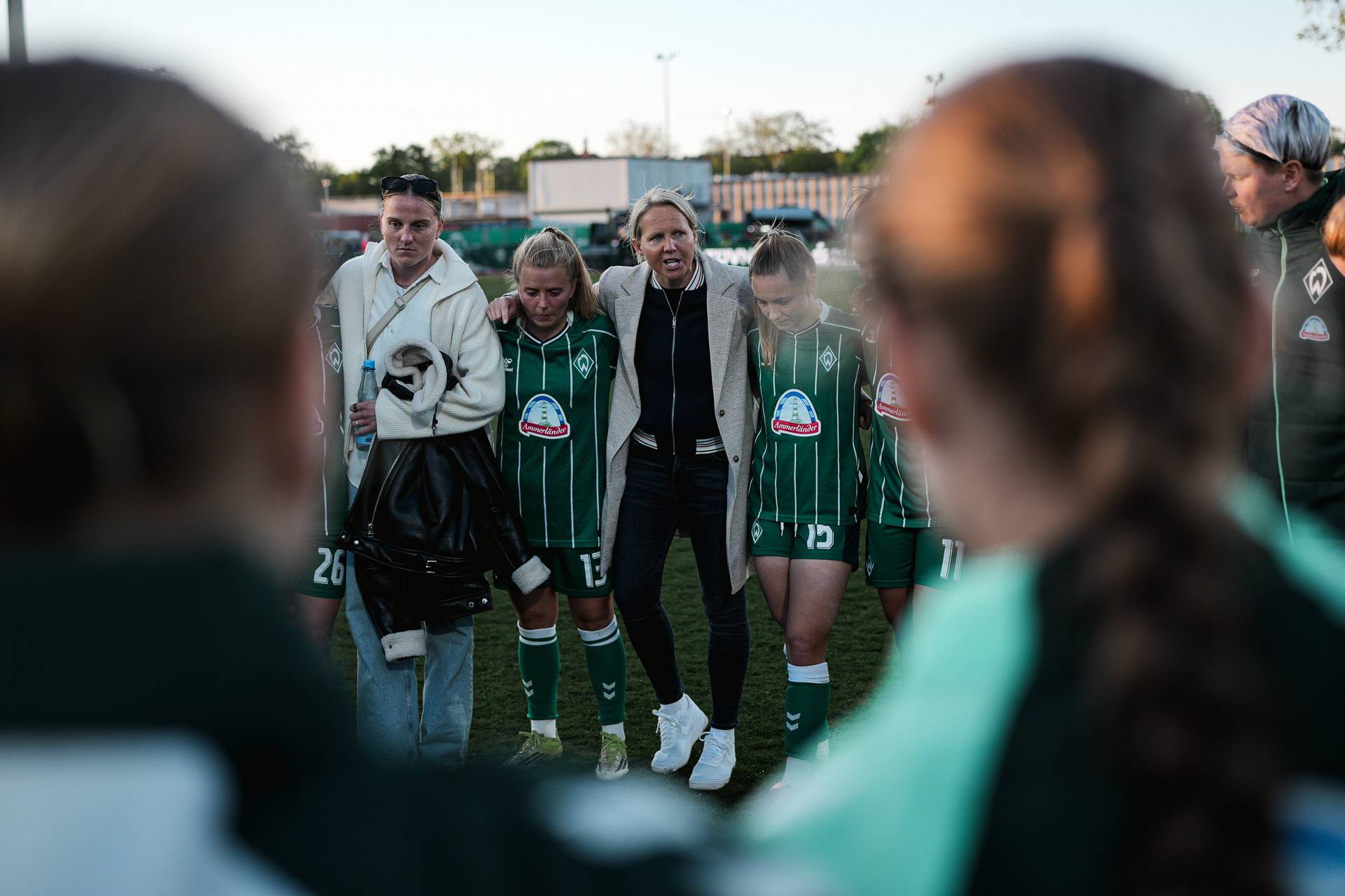 Eine Trainerin steht neben einer Frauenfußballmannschaft, die sich in grünen Trikots auf dem Spielfeld zusammengekauert hat.