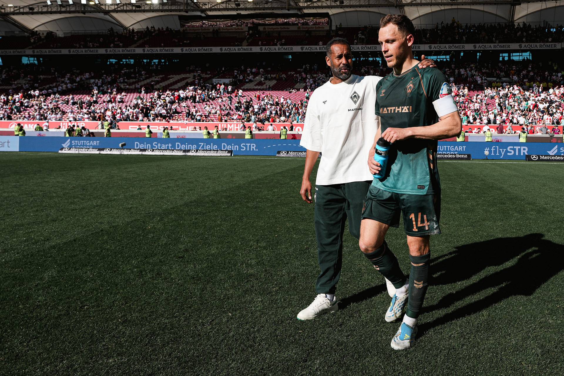 Zwei Fußballspieler verlassen das Spielfeld, einer in Spielkleidung mit einer blauen Flasche in der Hand, der andere in einem weißen Hemd. Im Hintergrund ist das Stadionpublikum zu sehen.