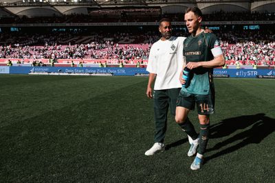 Zwei Fußballspieler verlassen das Spielfeld, einer in Spielkleidung mit einer blauen Flasche in der Hand, der andere in einem weißen Hemd. Im Hintergrund ist das Stadionpublikum zu sehen.