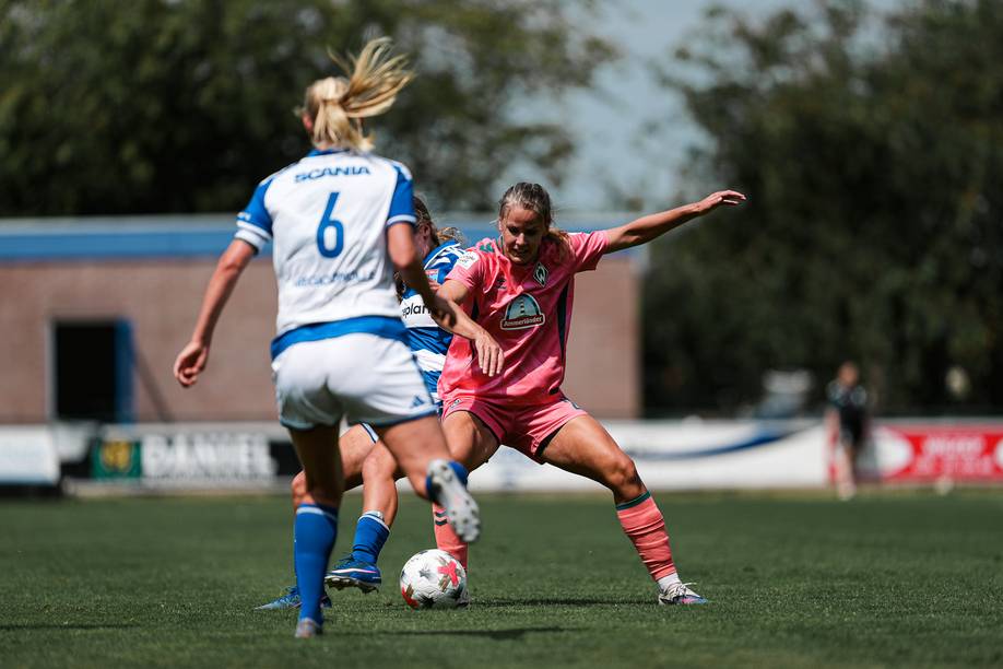 Zwei Fußballerinnen in Aktion; eine in Korallrot, die den Ball unter Kontrolle hat, steht einer anderen in einem weißen Trikot auf einem Rasenfeld gegenüber.