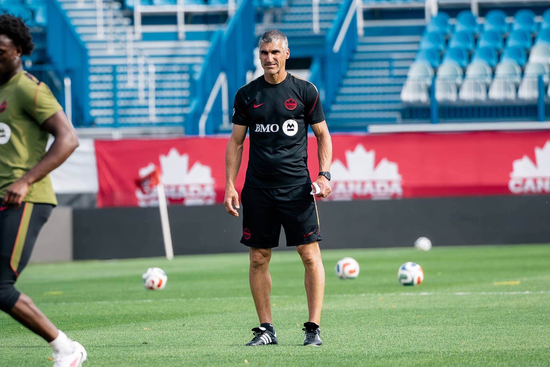 A coach in black training kit stands on a football pitch, with a Canada banner and empty seats visible in the background.