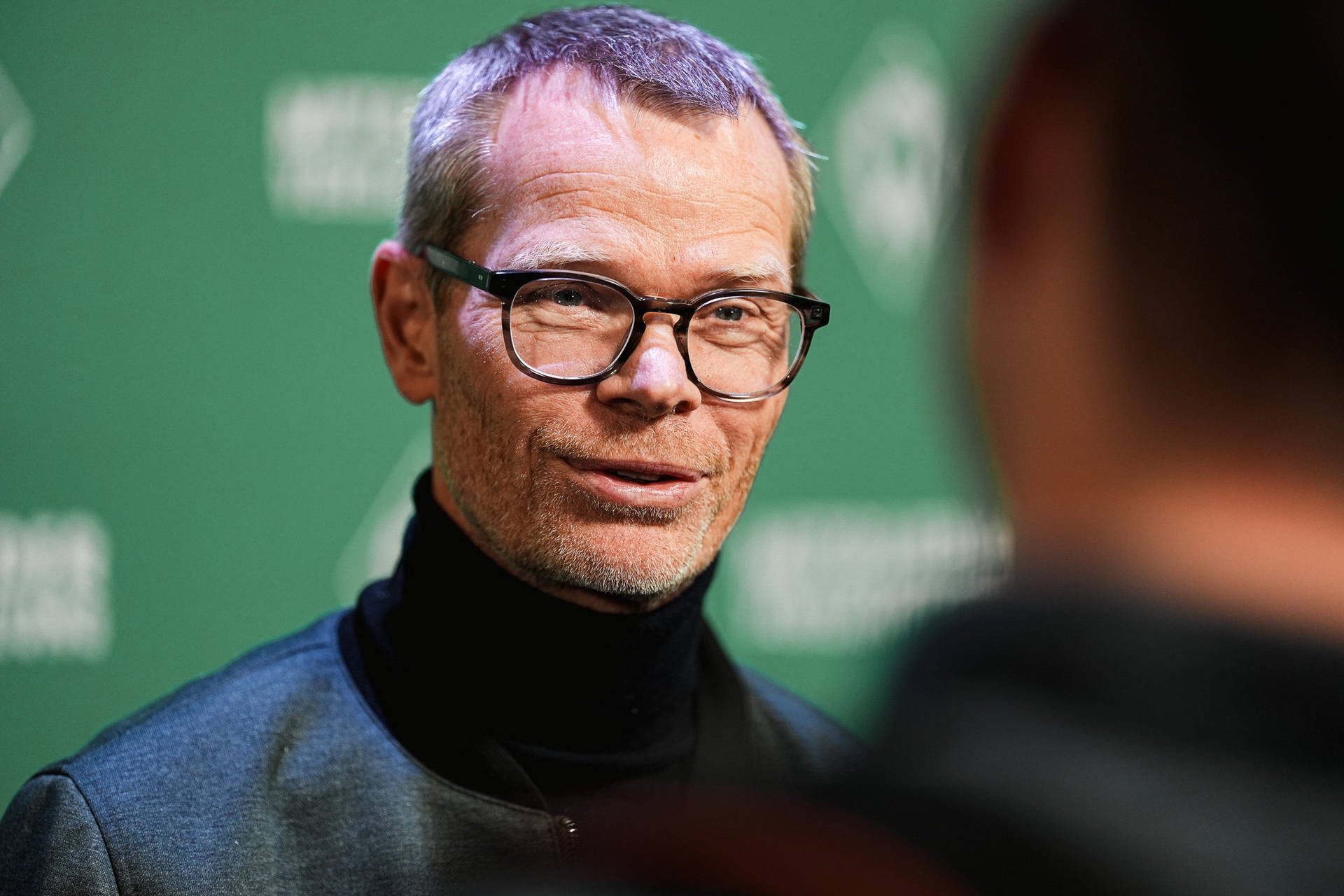 Mann mit Brille und kurzen Haaren spricht bei einer Veranstaltung, trägt eine dunkle Jacke und einen Rollkragenpullover; grüner Hintergrund mit weißem Text.