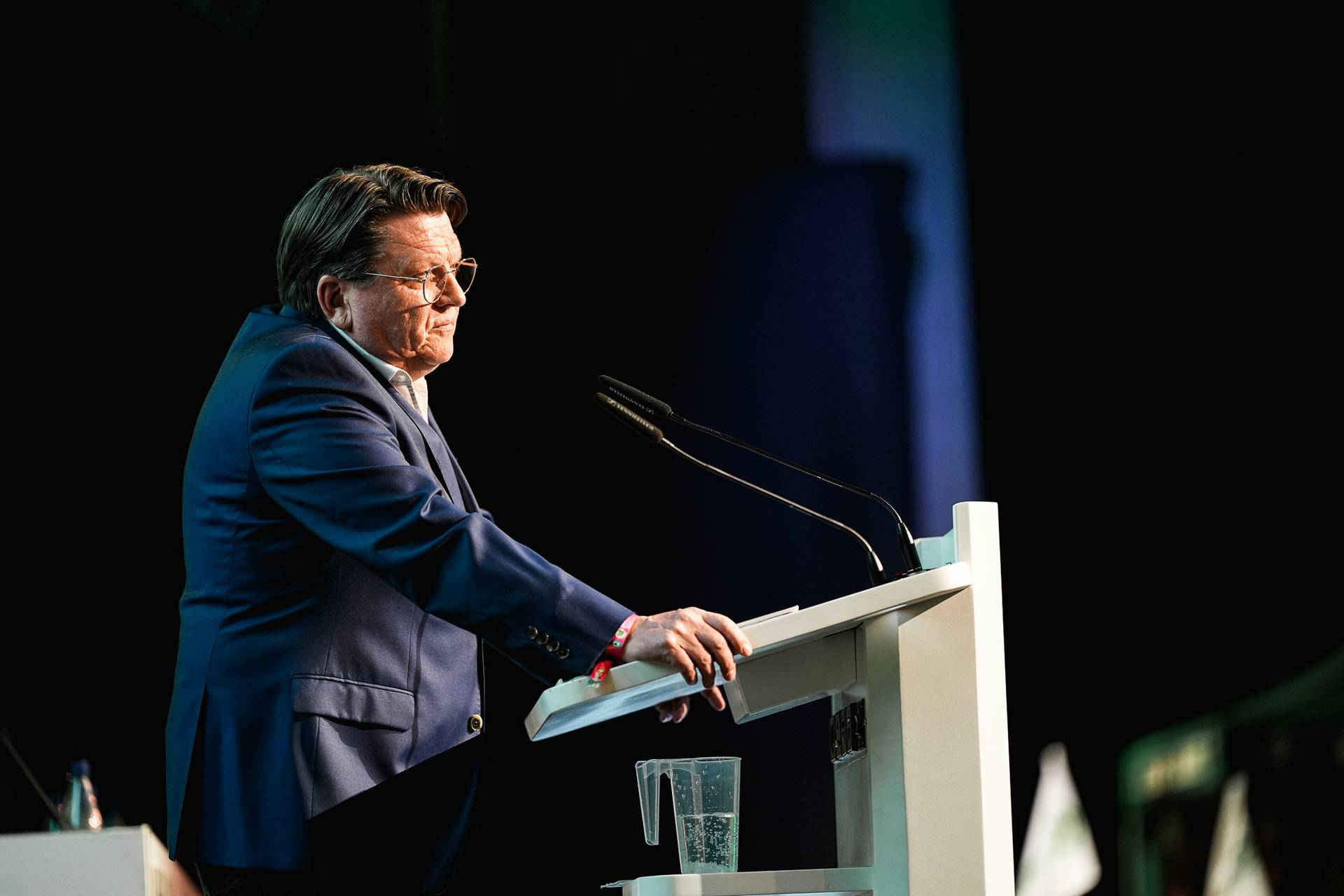 Mann in einem blauen Anzug spricht an einem Podium mit Mikrofonen vor einem dunklen Hintergrund.