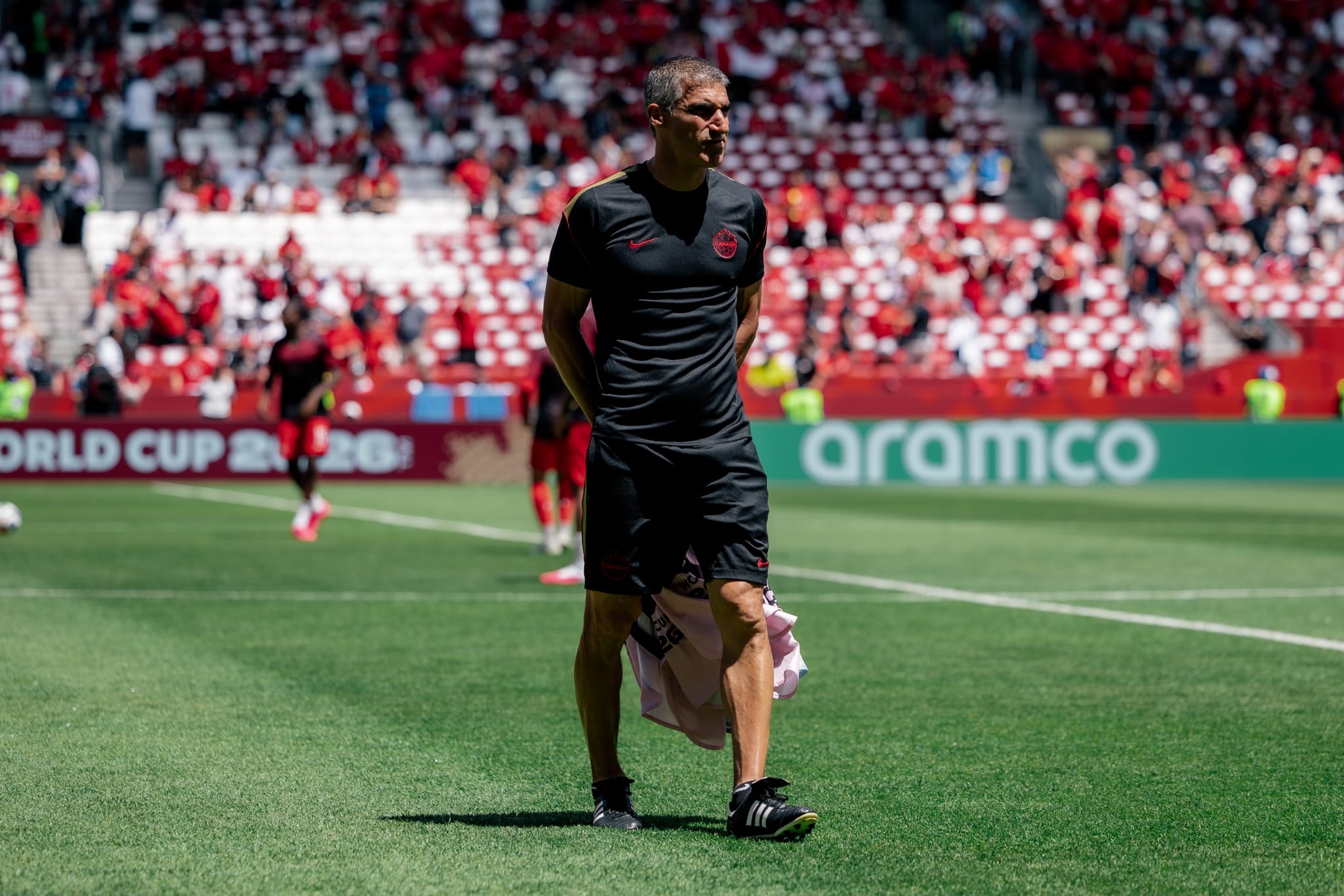 Paul Stalteri stands on the pitch in black training kit, holding a pink shirt, with supporters visible in the background.