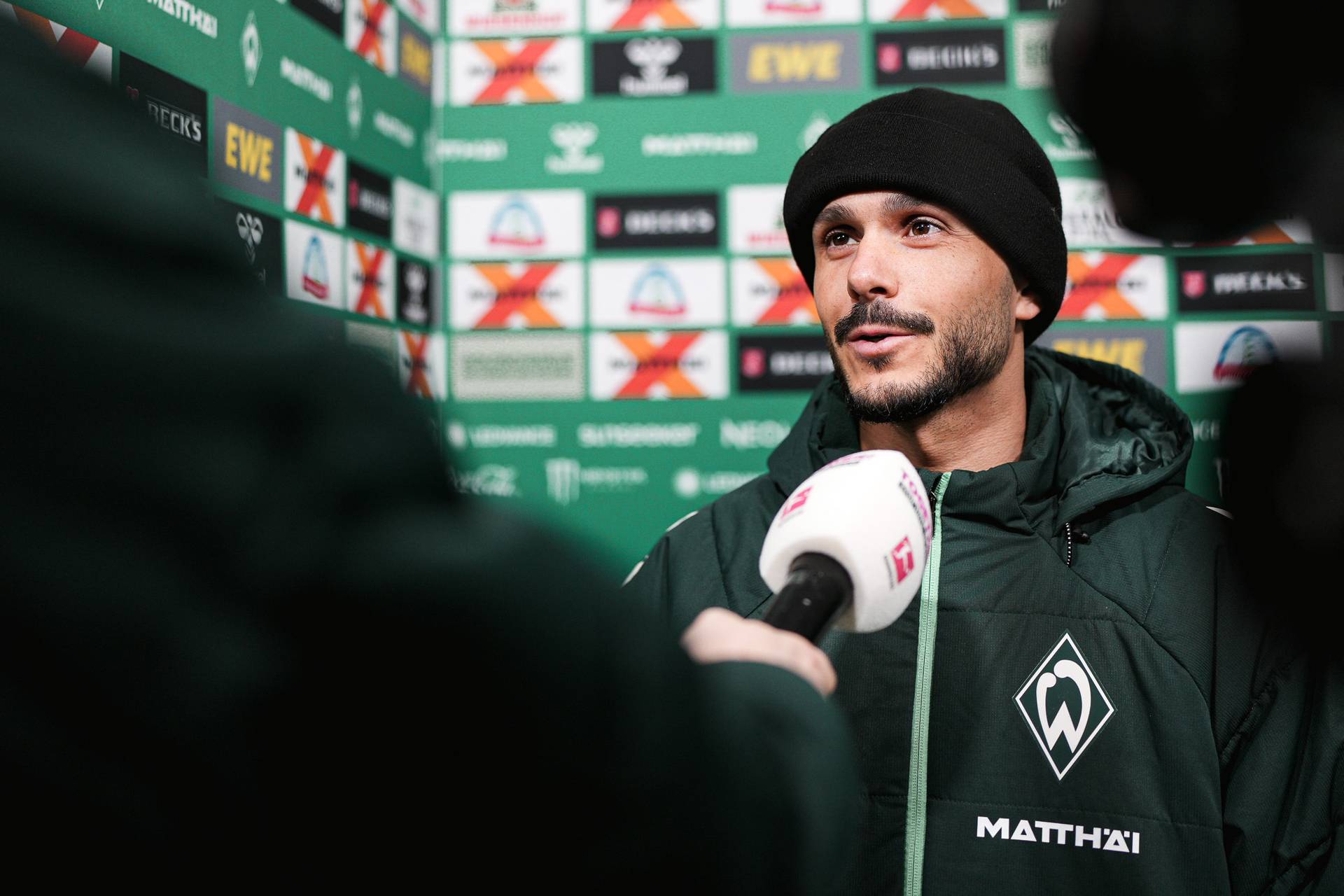 Ein Mann mit schwarzer Mütze und grüner Jacke spricht in Mikrofone; hinter ihm sind Logos und Werbeanzeigen auf einer Kulisse zu sehen.