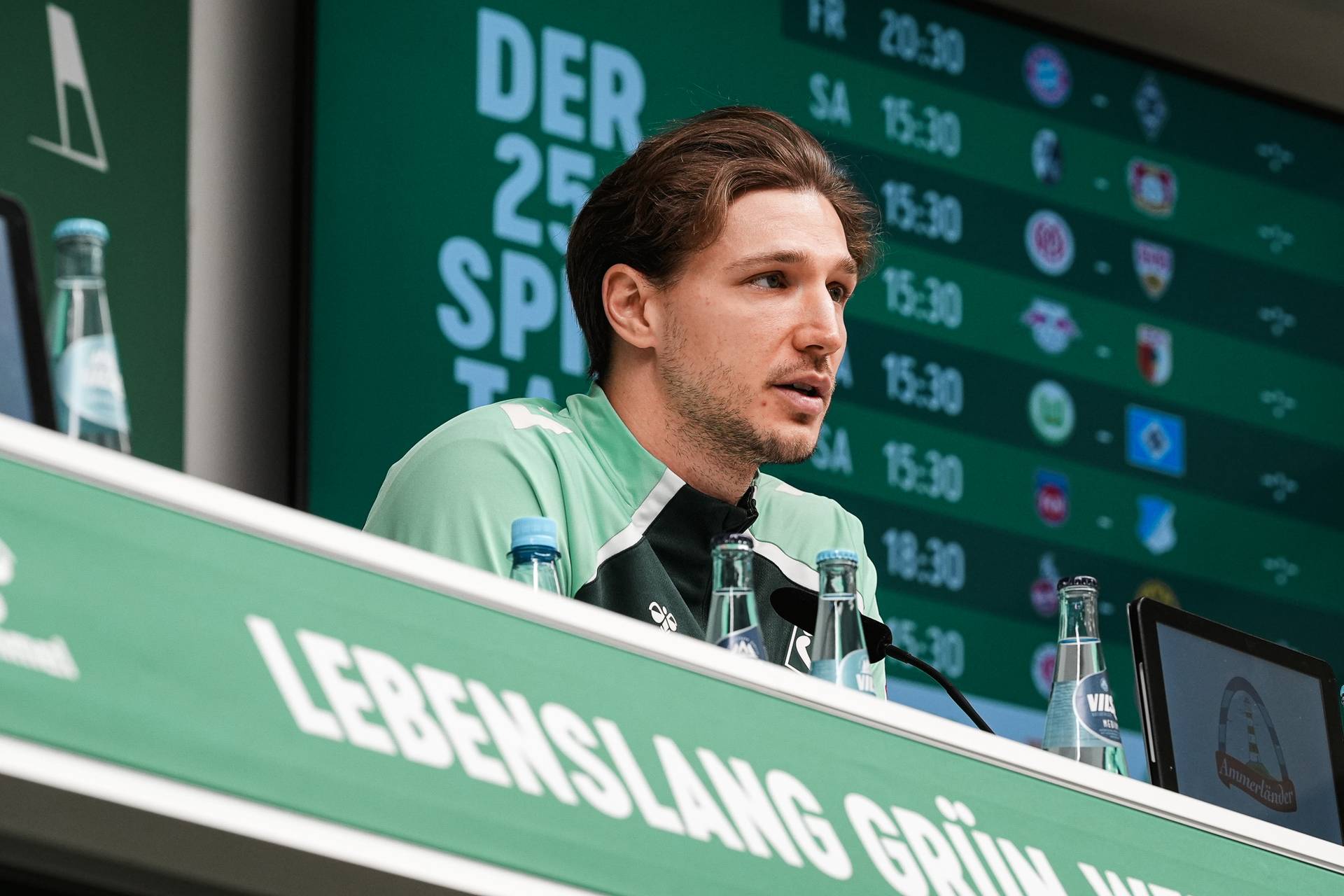 Eine Person in einer grünen Sportjacke spricht auf einer Pressekonferenz. Im Hintergrund ist ein Zeitplan zu sehen.