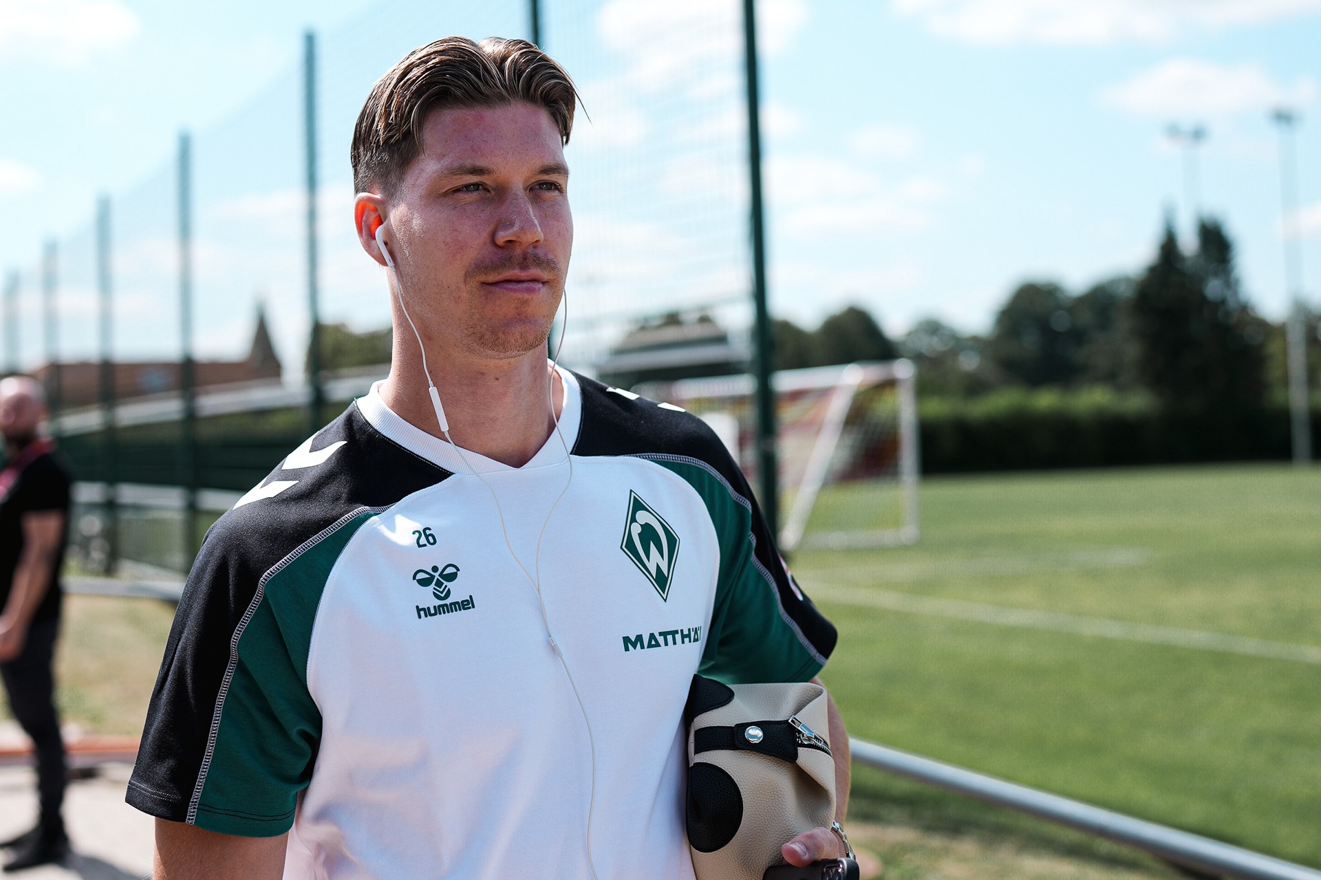 Ein Mann in Sportkleidung mit Kopfhörern, der eine Tasche in der Hand hält, geht bei strahlendem Himmel in der Nähe eines Fußballplatzes spazieren.