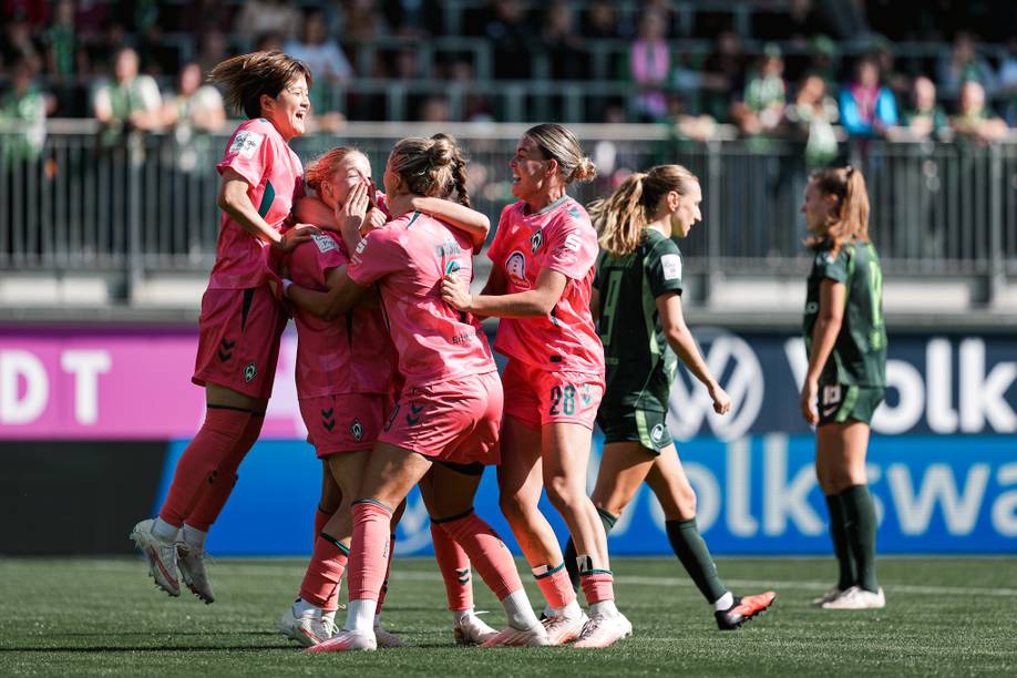 Fußballspieler in rosa Trikots feiern gemeinsam auf dem Spielfeld, während Spieler der gegnerischen Mannschaft in ihrer Nähe vorbeigehen.