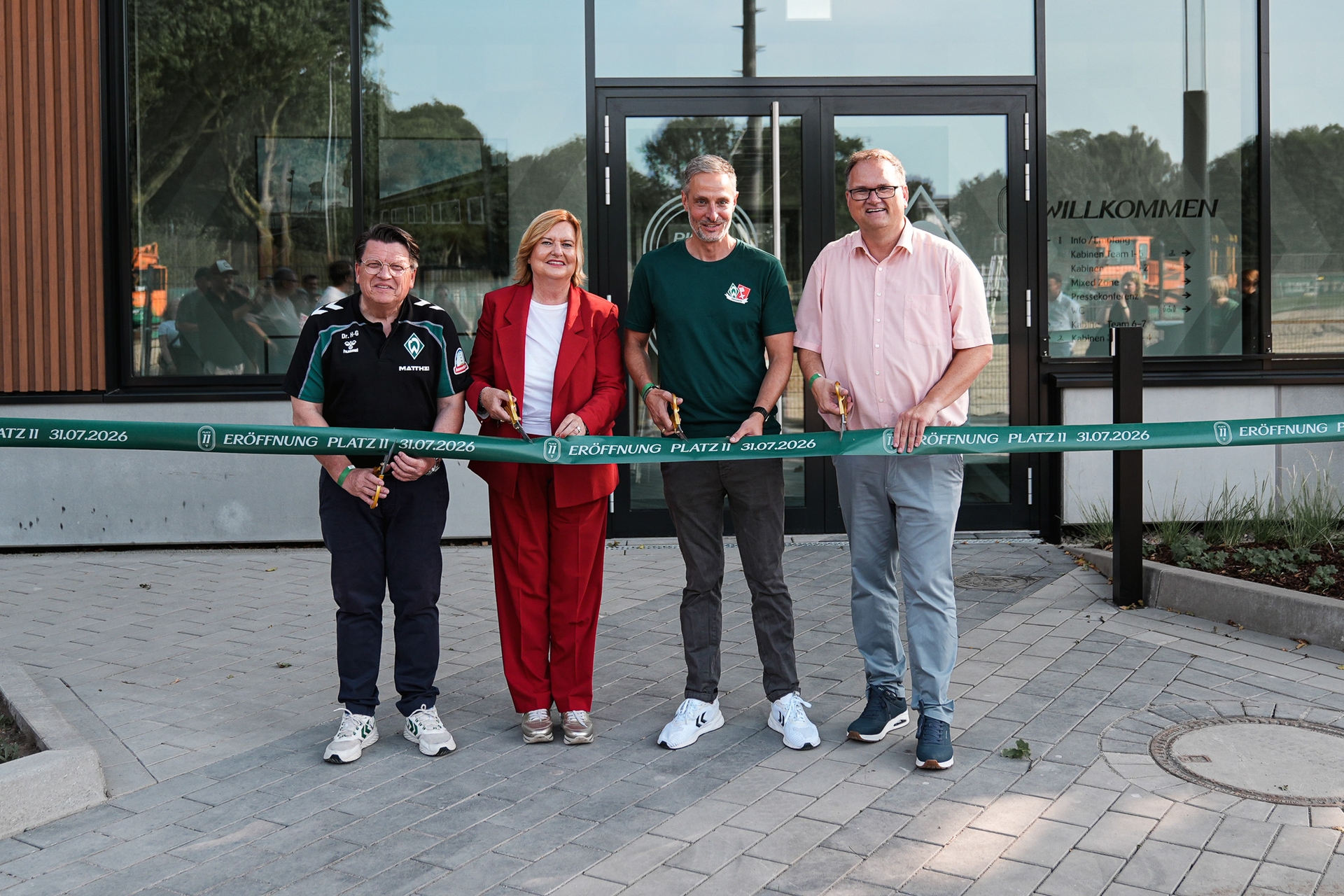 Vier Personen stehen vor einem Gebäude, halten Scheren in der Hand und durchtrennen ein grünes Band mit der Aufschrift „Eröffnung Platz II 31.07.2026“.