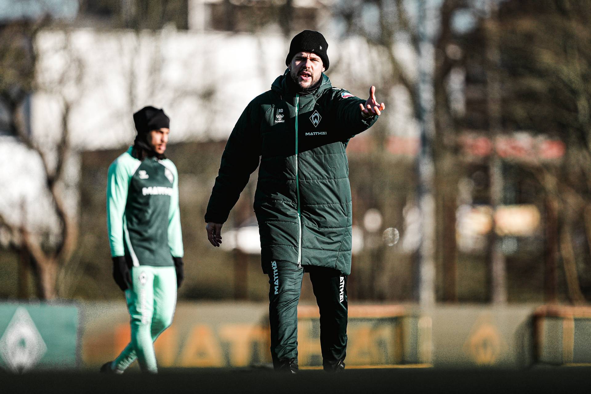 Ein Trainer in einer dunkelgrünen Jacke gestikuliert während eines Sporttrainings im Freien, im Hintergrund ist ein verschwommener Spieler zu sehen.