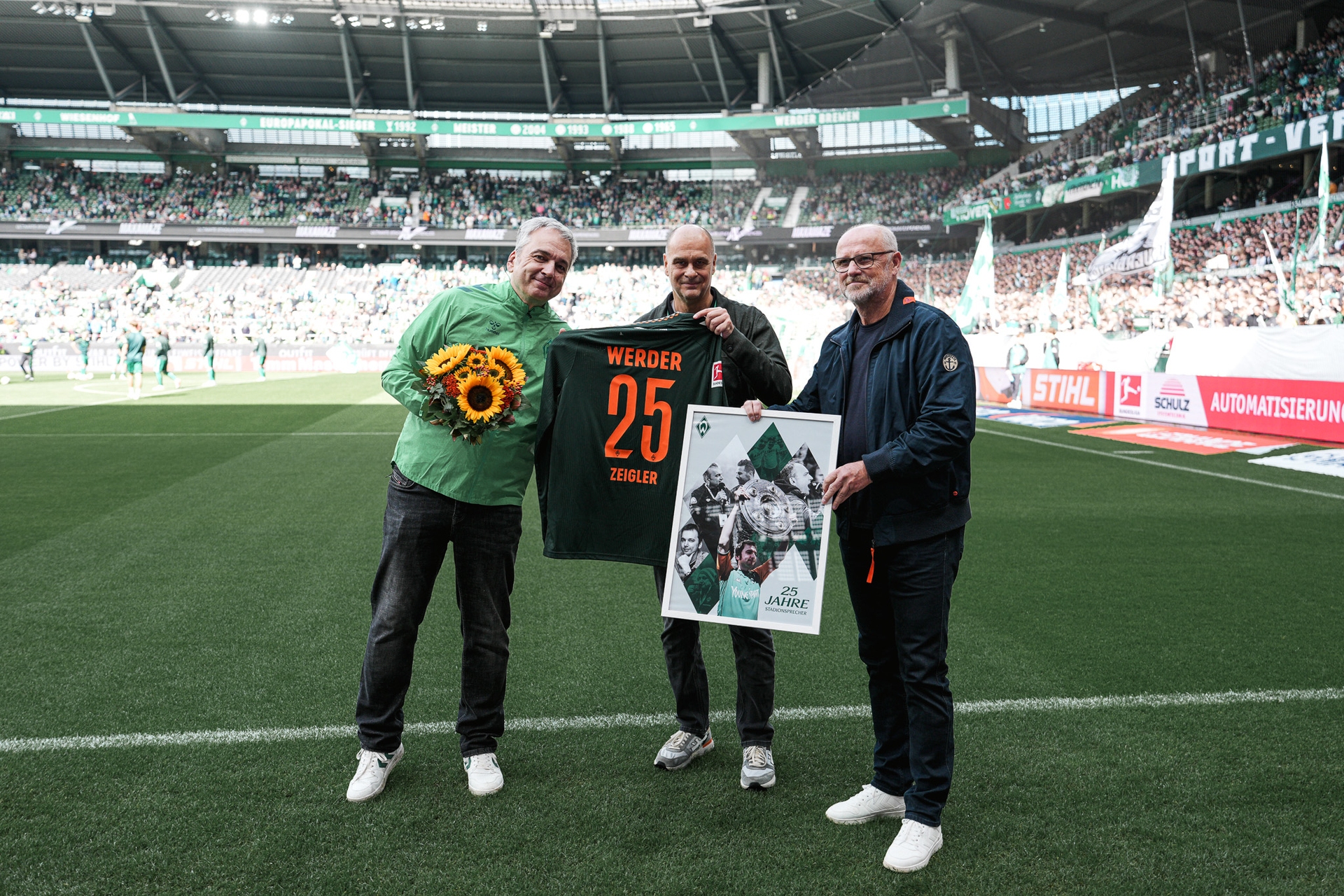 Drei Männer auf einem Fußballplatz halten einen Blumenstrauß, ein Trikot mit der Aufschrift „Werder 25“ und ein gerahmtes Foto in den Händen. Im Hintergrund füllt das Publikum das Stadion.
