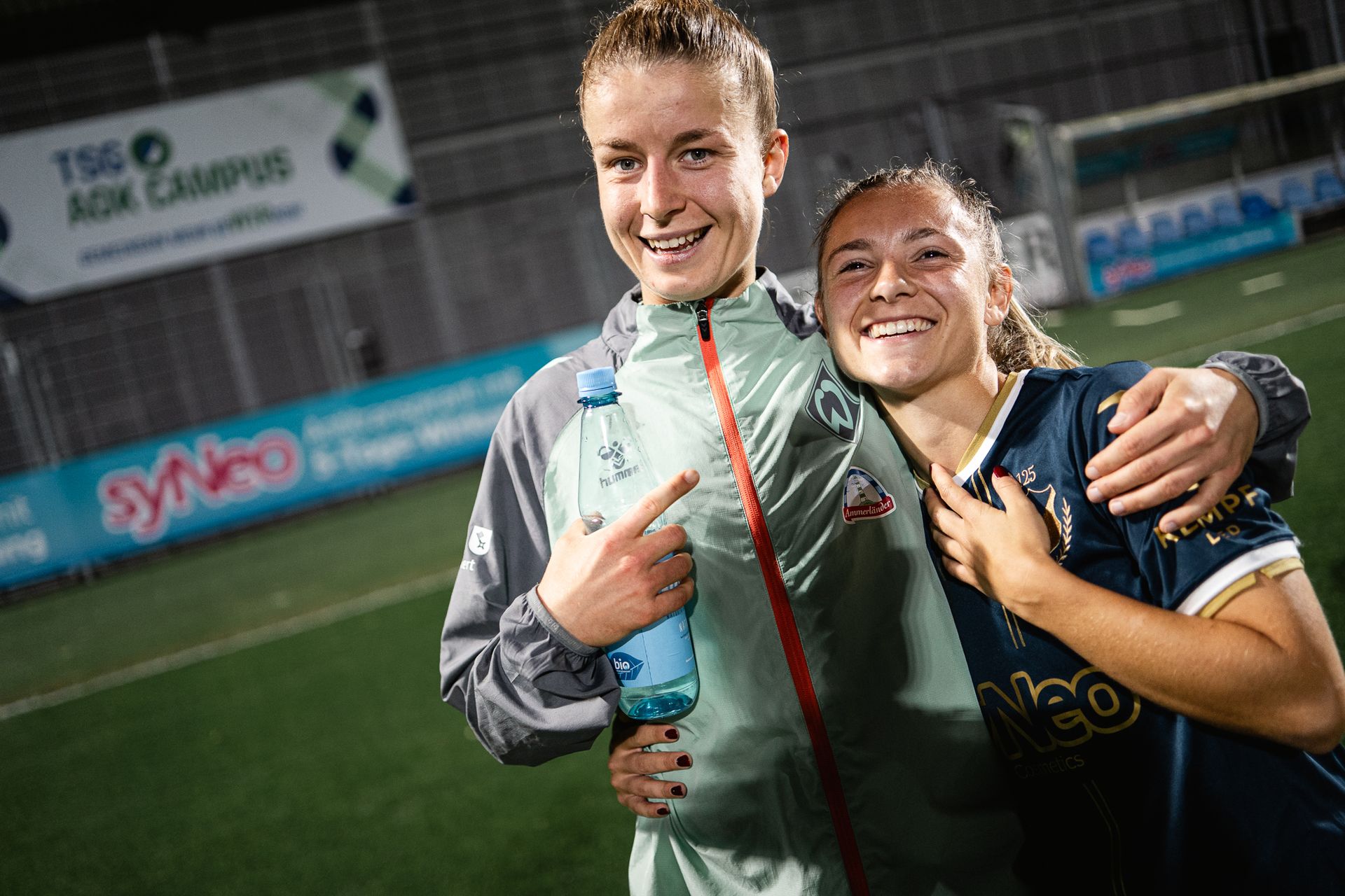 Michelle Ulbrich und Michelle Weiß lächeln in die Kamera in Sportbekleidung und stehen abends auf einem Fußballfeld, einer hält eine Wasserflasche in der Hand. Sie haben ihre Arme umeinander gelegt.
