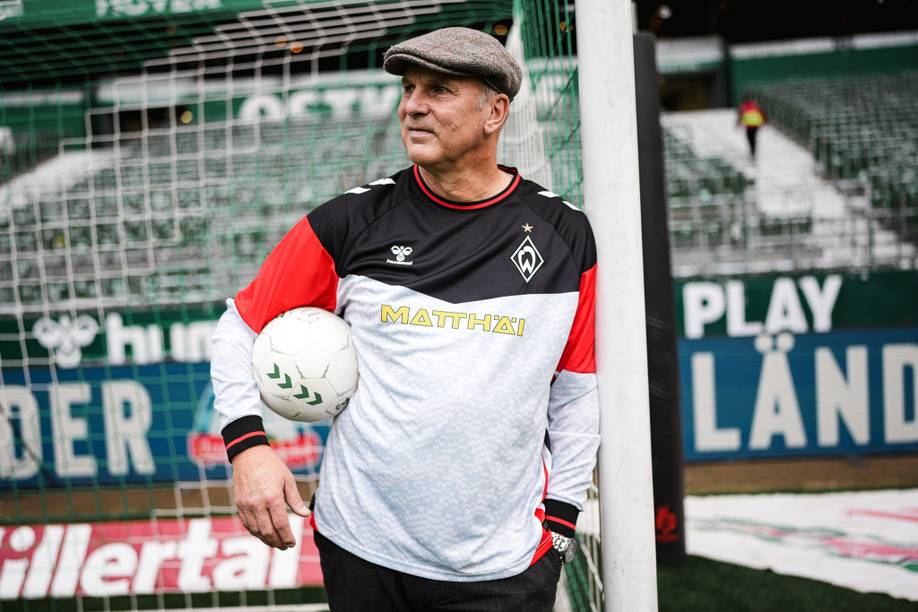 Oliver Reck trägt das Retro-Torwarttrikot. Er lehnt an einem Pfosten. Unter dem anderen Arm hat er einen Ball.