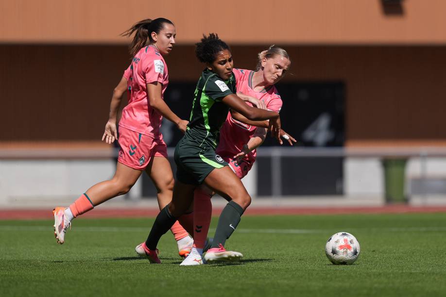Drei Fußballspielerinnen in Bewegung auf dem Spielfeld, zwei in rosa Trikots und eine in einem dunkelgrünen Trikot, die um den Ball kämpfen.