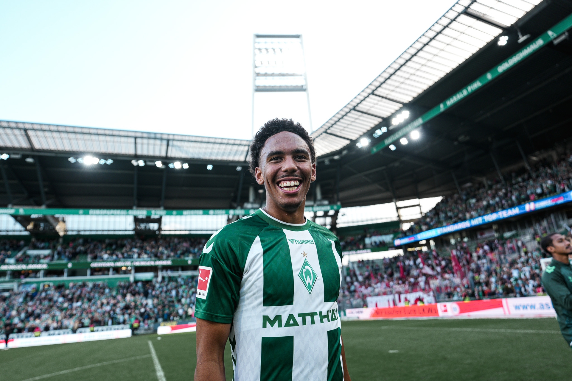 Ein Fußballspieler im grün-weißen Trikot grinst in die Kamera, er steht in einem Stadion. IM Hintergrund sind Flutlicht und Menschenmassen unscharf zu erkennen.
