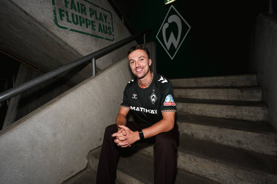 Eine Person sitzt auf einer Betontreppe, trägt ein schwarz-grünes Sporttrikot und lächelt. An der Wand dahinter sind ein „Fair Play“-Schild und ein Logo zu sehen.