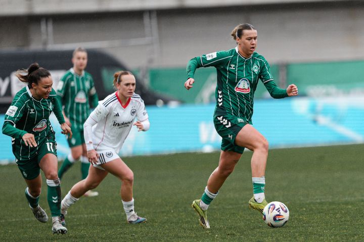 Eine Szene aus einem Fußballspiel, in der ein Spieler in grüner Spielkleidung den Ball dribbelt, während er auf dem Spielfeld von Gegnern in weißer und grüner Spielkleidung verfolgt wird.
