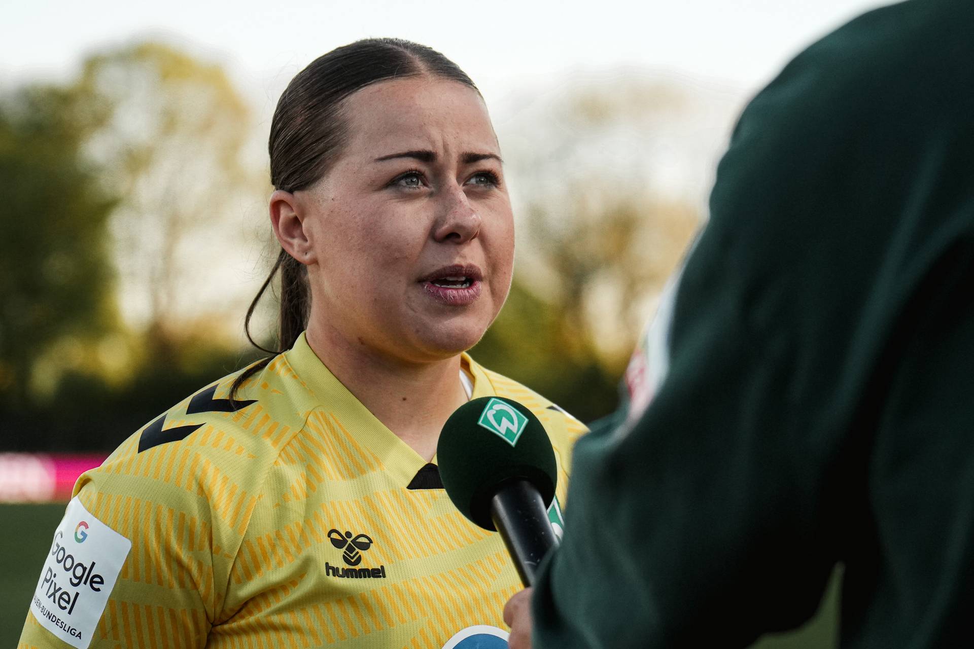 Eine Person in einem gelben Sporttrikot, die im Freien interviewt wird und ein grünes Mikrofon in der Hand hält, vor einem verschwommenen Hintergrund aus Bäumen.