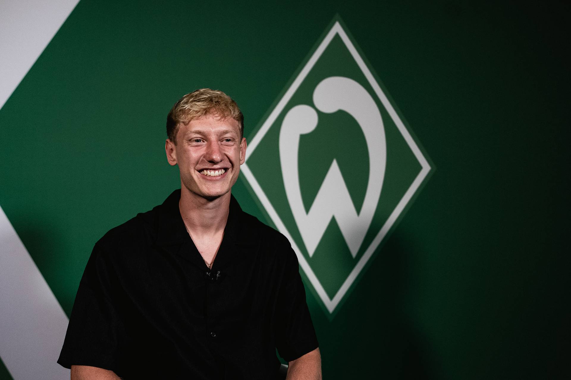 Oskar Wojcik lacht und trägt ein schwarzes Hemd. Hinter ihm ist eine Werder-Raute an der Wand.