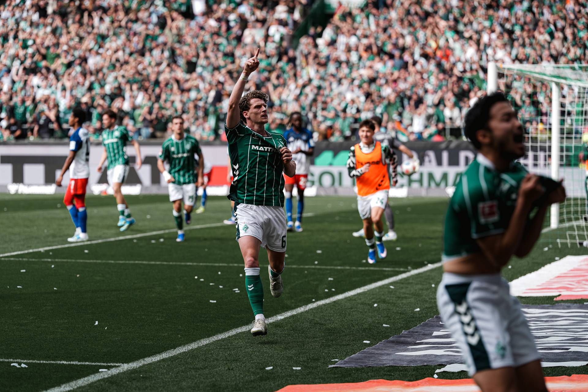 Ein Fußballspieler in grüner Kleidung feiert ein Tor, indem er mit dem Finger zum Himmel zeigt, während seine Mannschaftskameraden und die Zuschauer in einem vollbesetzten Stadion jubeln.