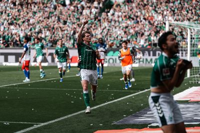 Ein Fußballspieler in grüner Kleidung feiert ein Tor, indem er mit dem Finger zum Himmel zeigt, während seine Mannschaftskameraden und die Zuschauer in einem vollbesetzten Stadion jubeln.