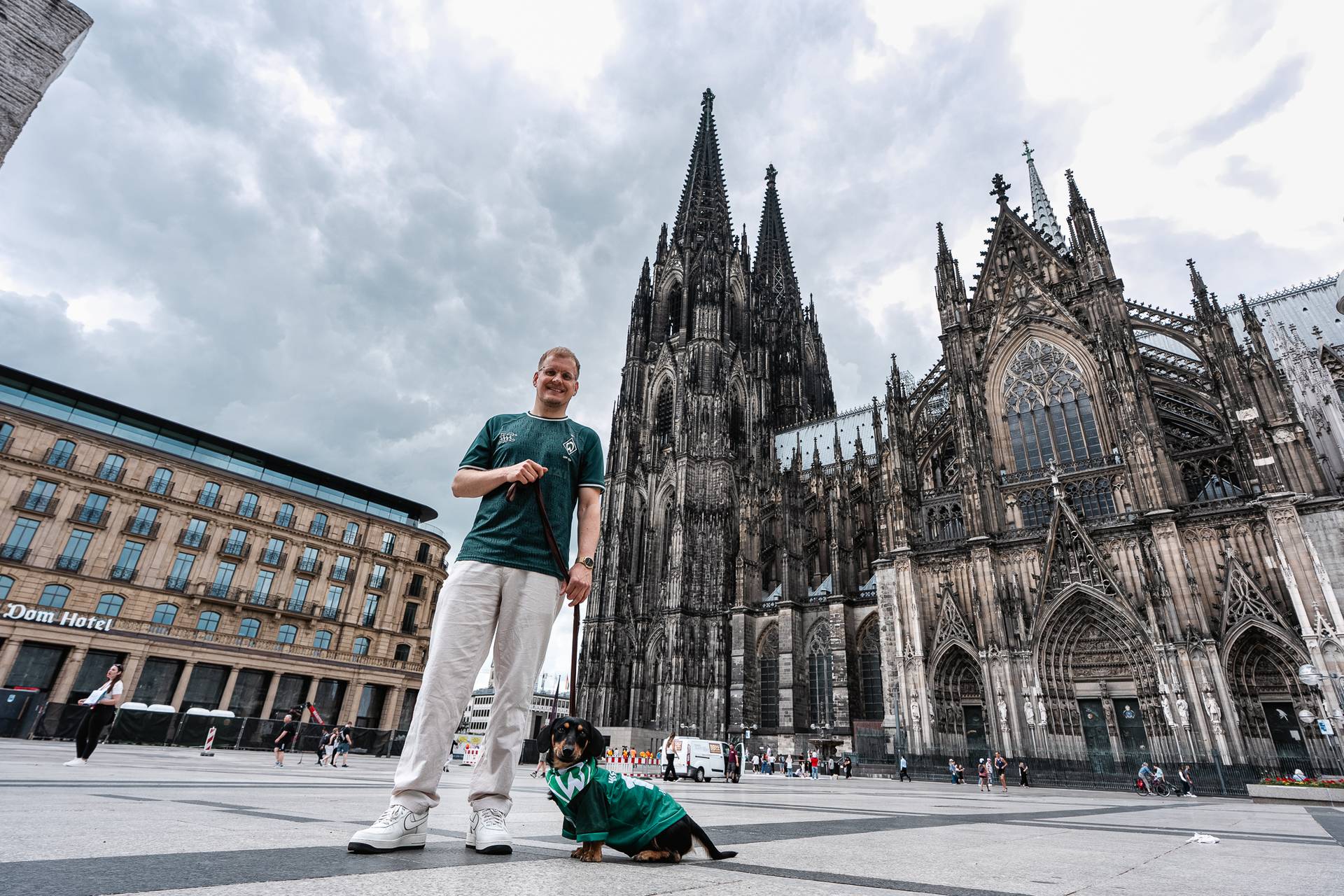 An einem bewölkten Tag steht ein Mann mit einem kleinen Hund vor dem Kölner Dom. Der Hund trägt ein grünes Werder-Trikot, und im Hintergrund ragt der Dom empor.